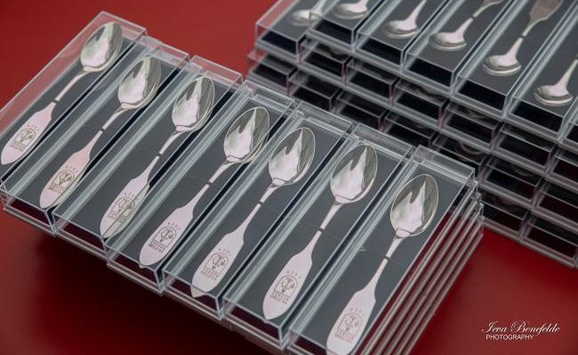 Iesaiņotas sudraba karotes, sakrautas rindās un kaudzēs uz sarkana fona. Katra karote atsevišķā caurspīdīgā plastmasas kastītē ar melnu ieliktni. Uz to rokturiem iegravēts brieža galvas simbols un teksts "SVEICAM VECĀKUS".