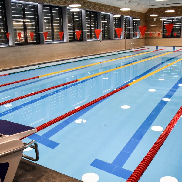 Kuldīgas peldbaseins. Iekštelpu peldbaseins ar gaiši zilu ūdeni, vairākām zilām celiņu līnijām un sarkani dzeltenām celiņu virvēm. Priekšplānā redzams starta bloks ar numuru "1", bet virs baseina izkārtoti sarkani karodziņi.