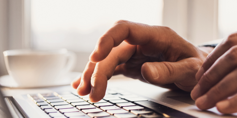 Rokas drukā uz klēpjdatora tastatūras, fonā kafijas tase pie gaiša loga.