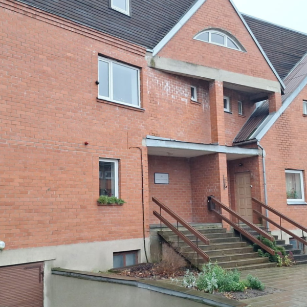 Sarkana ķieģeļu ēka ar stāviem jumtiem un dažādu formu logiem. Galvenā ieeja ar koka margām ved uz durvīm zem nelielas nojumes.