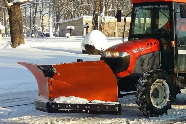 Sarkans traktors ar oranžu sniega lāpstu stāv uz sniegotas ceļa malas saulainā ziemas dienā.
