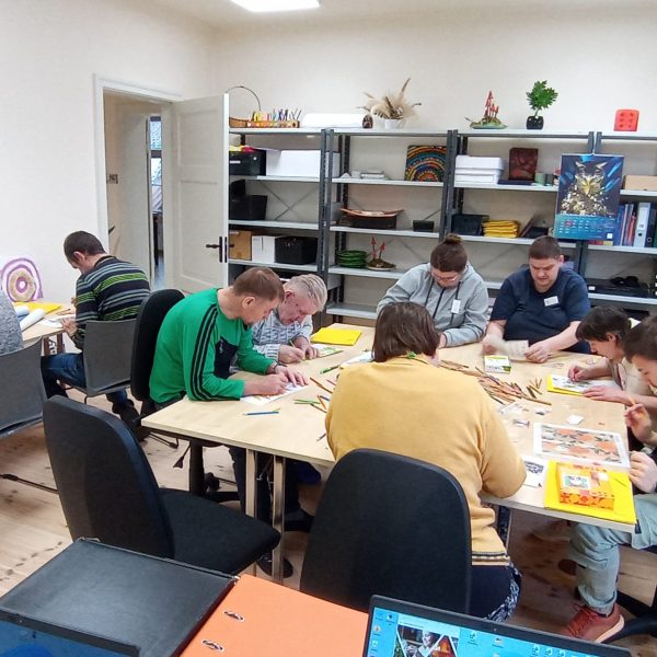 Vairāki cilvēki sēž pie diviem galdiem un ir iesaistīti radošā darbnīcā, zīmējot un krāsojot ar krāsu zīmuļiem. Fonā redzams plaukts ar grāmatām, mapēm un dažādiem dekoratīviem priekšmetiem, ieskaitot plakātu ar fantastiskas radības attēlu.