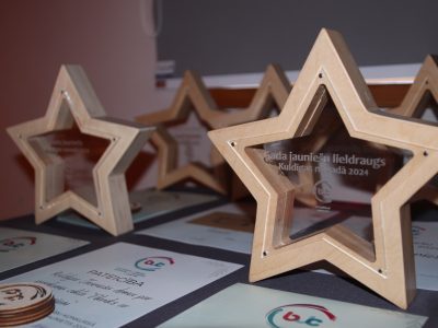 Vairāki koka zvaigznes formas apbalvojumi ar caurspīdīgiem centriem un sertifikāti uz galda. Uz priekšējā apbalvojuma ir uzraksts: "Gada jauniešu lieldraugs Kuldīgas novadā 2024".