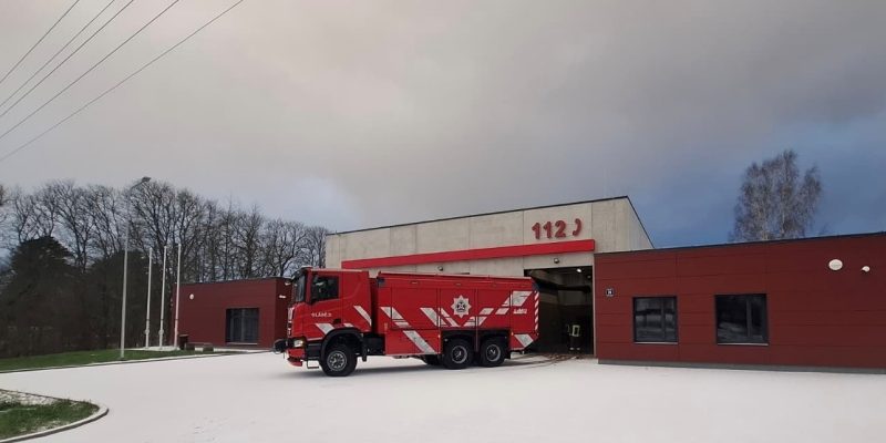 Sarkana ugunsdzēsēju mašīna ar baltām svītrām, uzrakstu "BLAŅES", emblēmu un numuru "402" novietota pie moderna ugunsdzēsēju depo ēkas, uz kuras fasādes redzami lieli sarkani cipari "112". Zeme ir apsnigusi.