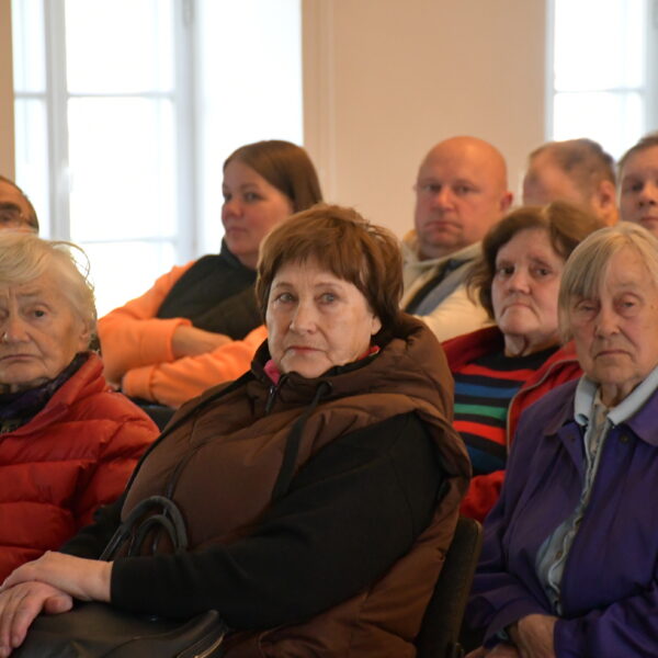 Cilvēku grupa, galvenokārt gados vecāki cilvēki, sēž iekštelpās, klausoties vai vērojot priekšā notiekošo. Priekšplānā redzamas trīs gados vecākas sievietes: viena ar baltiem matiem sarkanā jakā, otra ar brūniem matiem brūnā vestē, trešā ar sirmiem matiem violetā jakā.