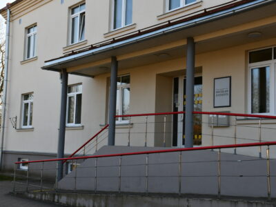 Gaišas krāsas ēka ar uzbrauktuvi un sarkaniem margām pie ieejas, virs kuras ir zīme ar uzrakstu "SOCIĀLAIS DIENESTS". Pa kreisi plīvo Latvijas karogs.