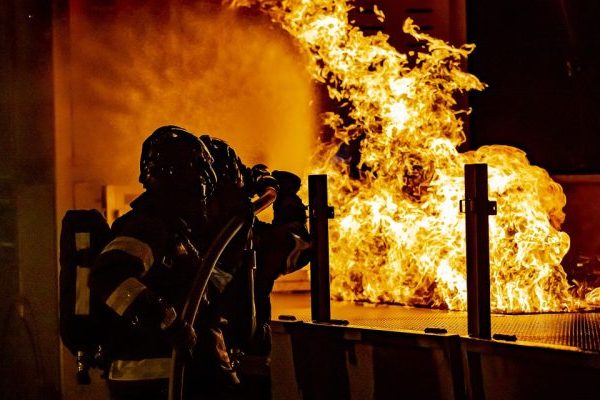 Divi ugunsdzēsēji aizsargtērpos dzēš lielu, spilgtu ugunsgrēku tumšā telpā, viens tur ugunsdzēsības šļūtenes uzgali.