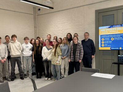 Aptuveni divdesmit jaunieši un viens pieaugušais vīrietis pozē grupas fotogrāfijai gaišā telpā. Aiz viņiem ir liels ekrāns ar informāciju par "JAUNO SPECIĀLISTU SKOLU" un "2. nodarbība – būvvaldes un nekustamo īpašumu speciālisti". Teksts uz ekrāna min: "OTRDIEN 27. janvārī", "No plkst. 13.00 līdz plkst. 17.00", "Vieta: Adatu fabrika", "Mērķauditorija: 8.-12. klases skolēni" un "PIESAKIES ieeja.lv/tilts-s2".