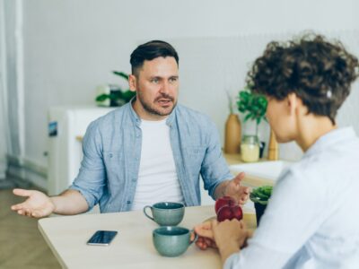 Vīrietis gaiši zilā kreklā, sēžot pie galda, runā ar sievieti, kas sēž viņam pretī, un izteiksmīgi žestikulē ar rokām. Uz gaišā koka galda ir krūzes, sarkani āboli un viedtālrunis.