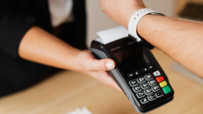 Tuvplānā redzams, kā cilvēks ar baltu viedpulksteni uz rokas veic bezkontakta maksājumu, pieliekot to pie melna POS termināļa, ko tur cits cilvēks.