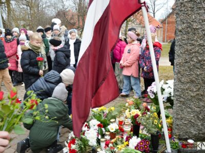 Bērni noliek ziedus un iedegtas sveces pie akmens pieminekļa, kura pakājē jau izvietoti daudzi ziedi un svecītes. Blakus plīvo Latvijas karogs. Fonā stāv vēl citi bērni un pieaugušie. Komunistiskā genocīda upuru piemiņas pasākumi novadā
