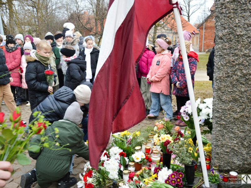 Bērni noliek ziedus un iedegtas sveces pie akmens pieminekļa, kura pakājē jau izvietoti daudzi ziedi un svecītes. Blakus plīvo Latvijas karogs. Fonā stāv vēl citi bērni un pieaugušie. Komunistiskā genocīda upuru piemiņas pasākumi novadā