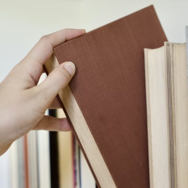 Cilvēka roka izvelk brūnu grāmatu no grāmatu plaukta, kurā redzamas arī citas grāmatas.