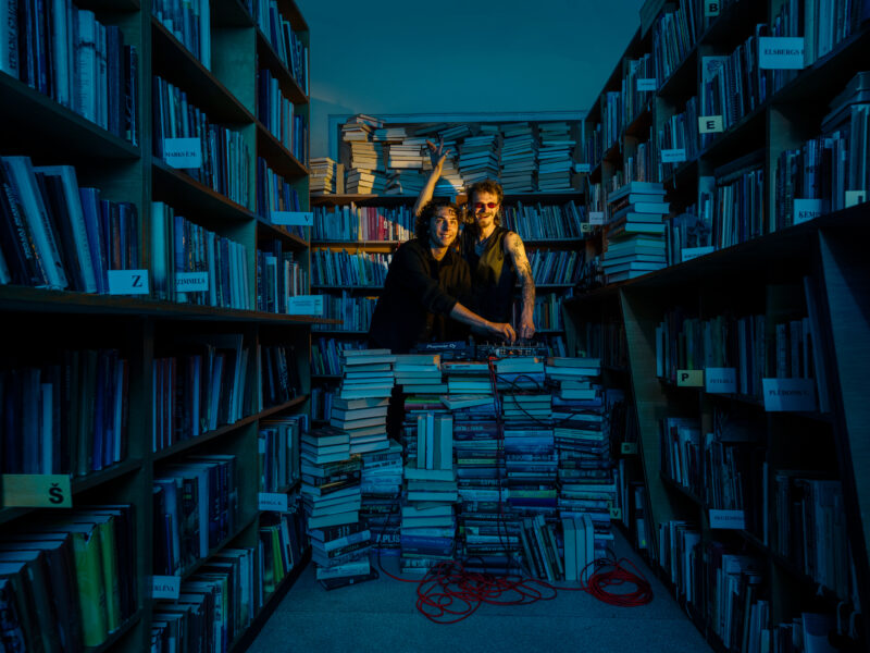 Divi smaidīgi vīrieši, viens ar tumšām saulesbrillēm un tetovētām rokām, blāvi apgaismotā bibliotēkā veido mūziku pie dīdžeja aprīkojuma "Pioneer DJ", kas novietots uz augstas grāmatu kaudzes. Viņu apkārtnē ir plaukti pilni ar grāmatām un grīda ir nokrauta ar papildu grāmatu kaudzēm.